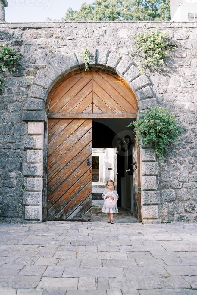 Little girl with a pink rabbit comes out of the arched wooden doors into the courtyard of an ancient castle photo