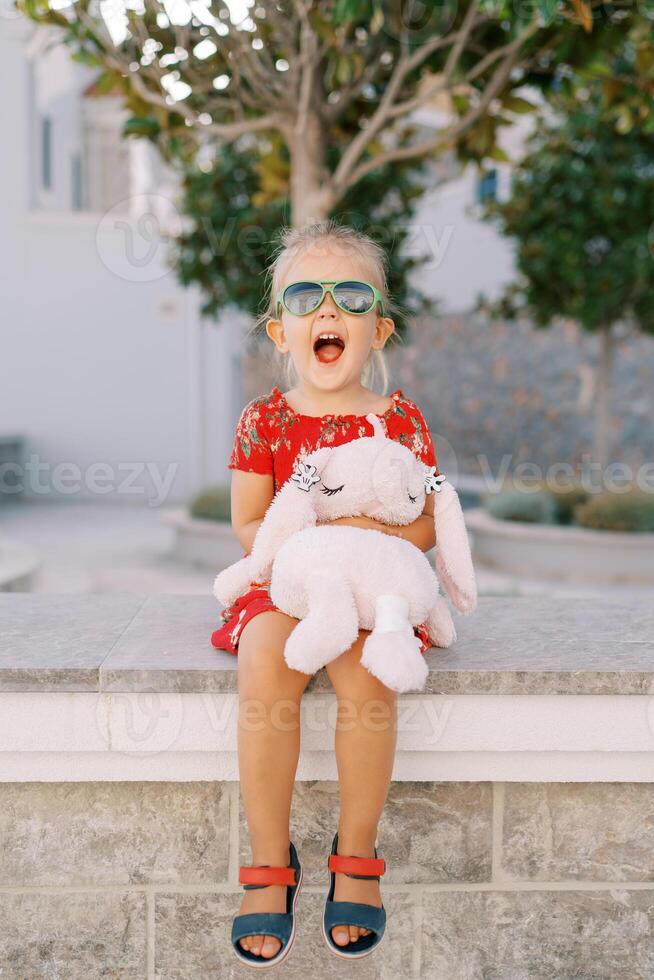 Little screaming girl sits hugging a soft toy rabbit on a stone fence in the garden photo