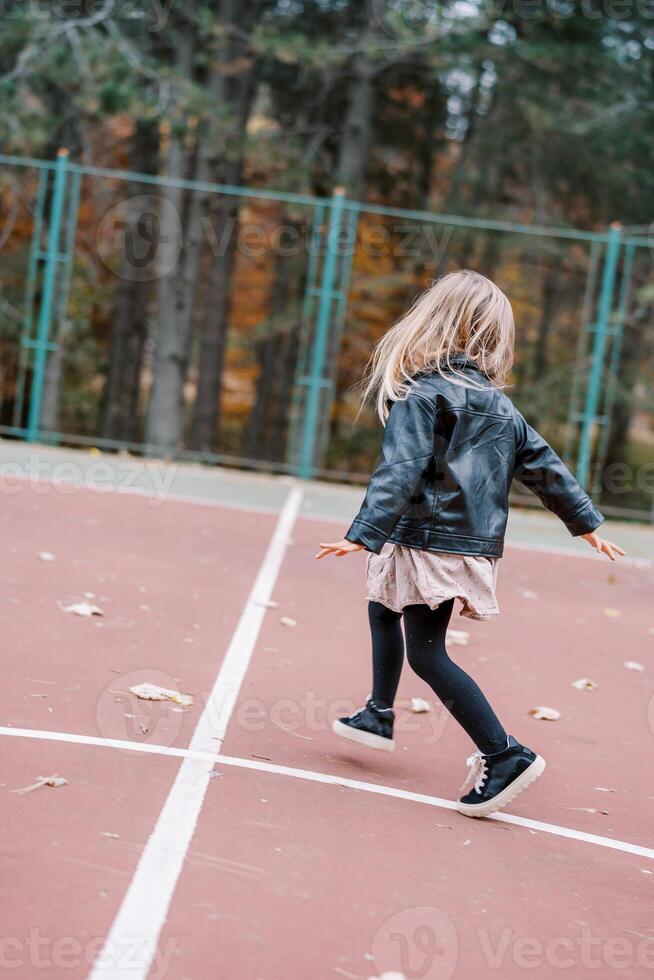 Little girl is skipping along the playground, looking to the side. Back view photo