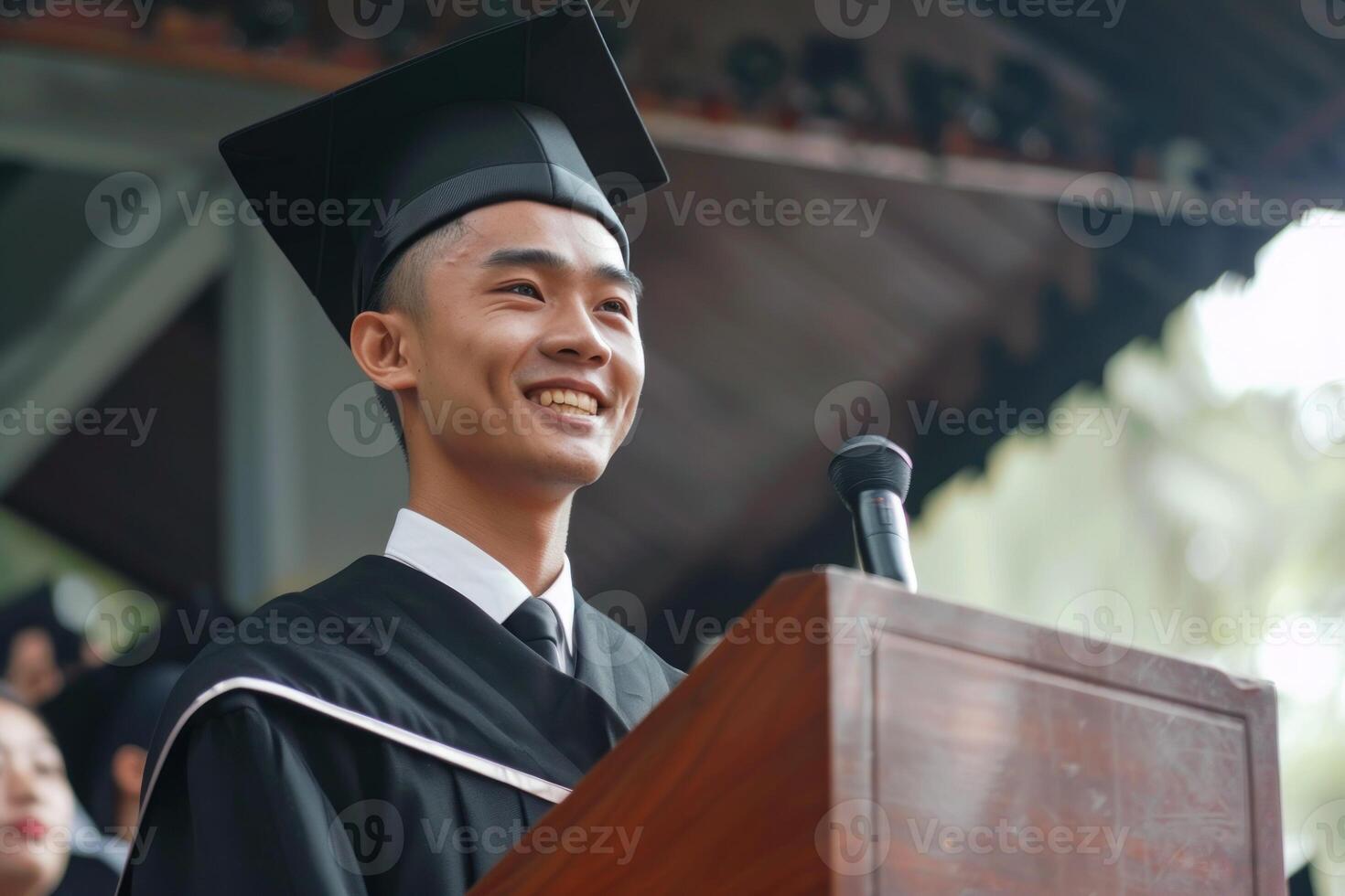 A man in a black graduation robe stands in front of a microphone photo