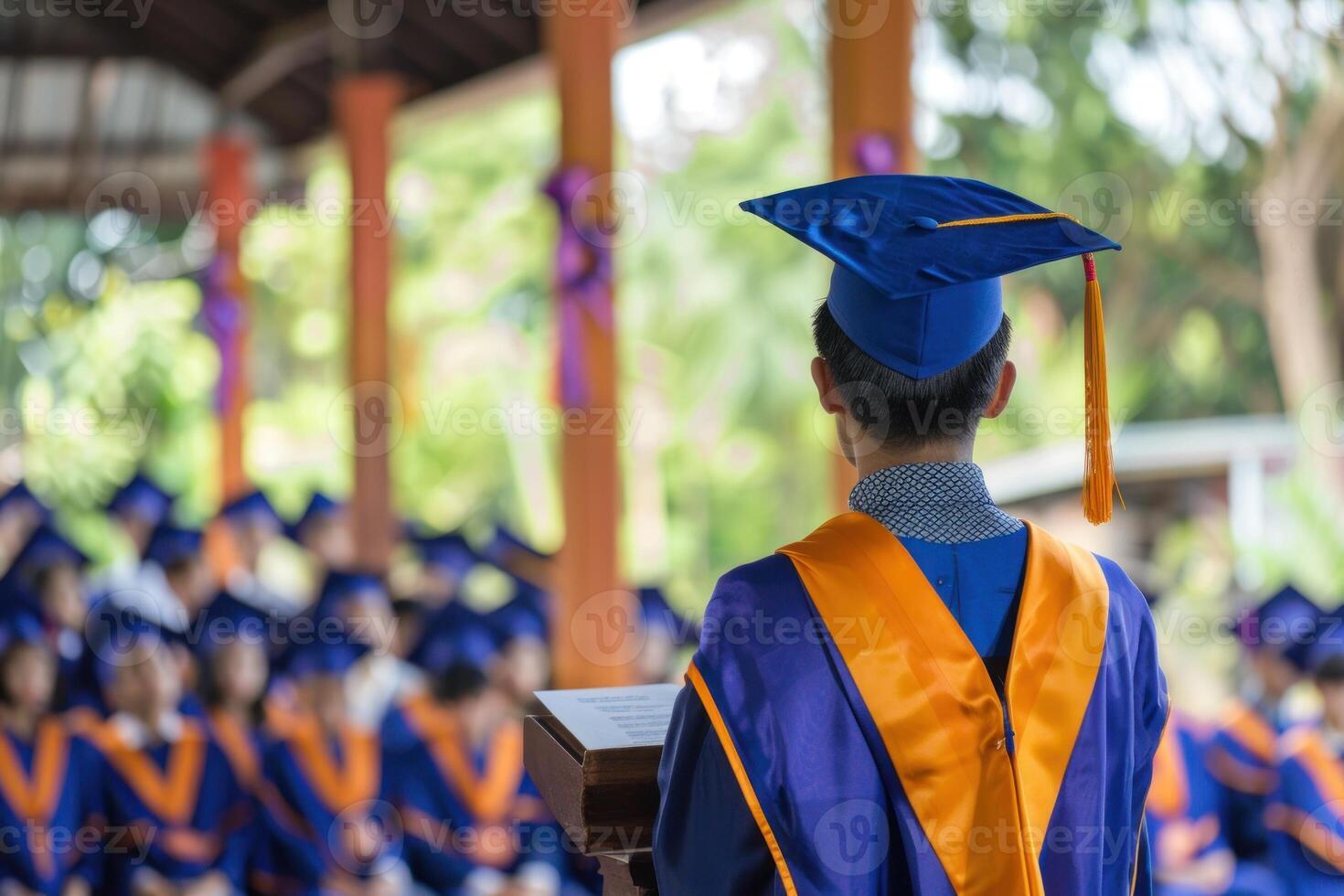 A man in a black graduation robe stands in front of a microphone photo