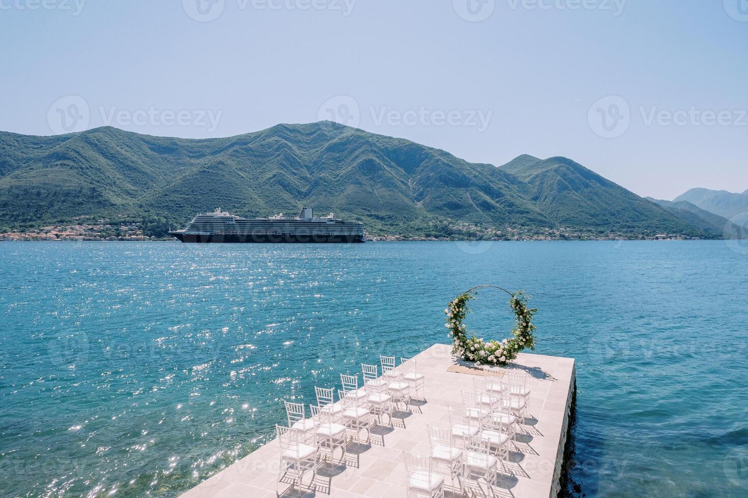 Rows of white chairs stand in front of a round wedding arch by the sea with a passing cruise ship in the background photo