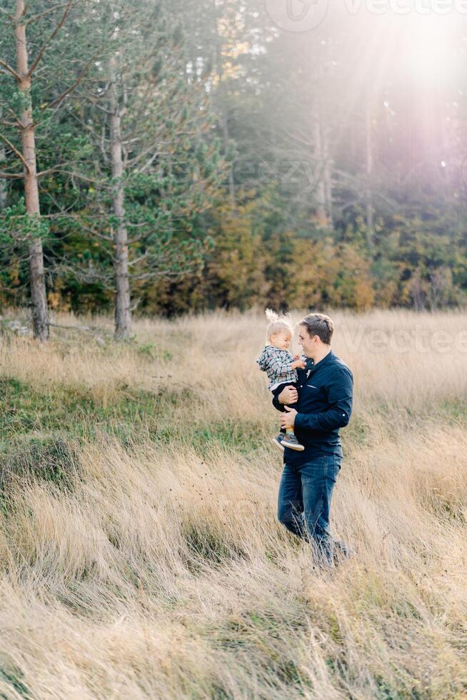 papá con un pequeño niña en su brazos camina mediante el alto césped a el borde de el bosque foto