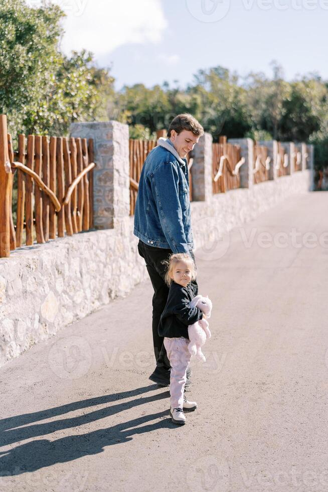 Little girl looks back while walking with her dad holding hands along the road along the fence. Back view photo