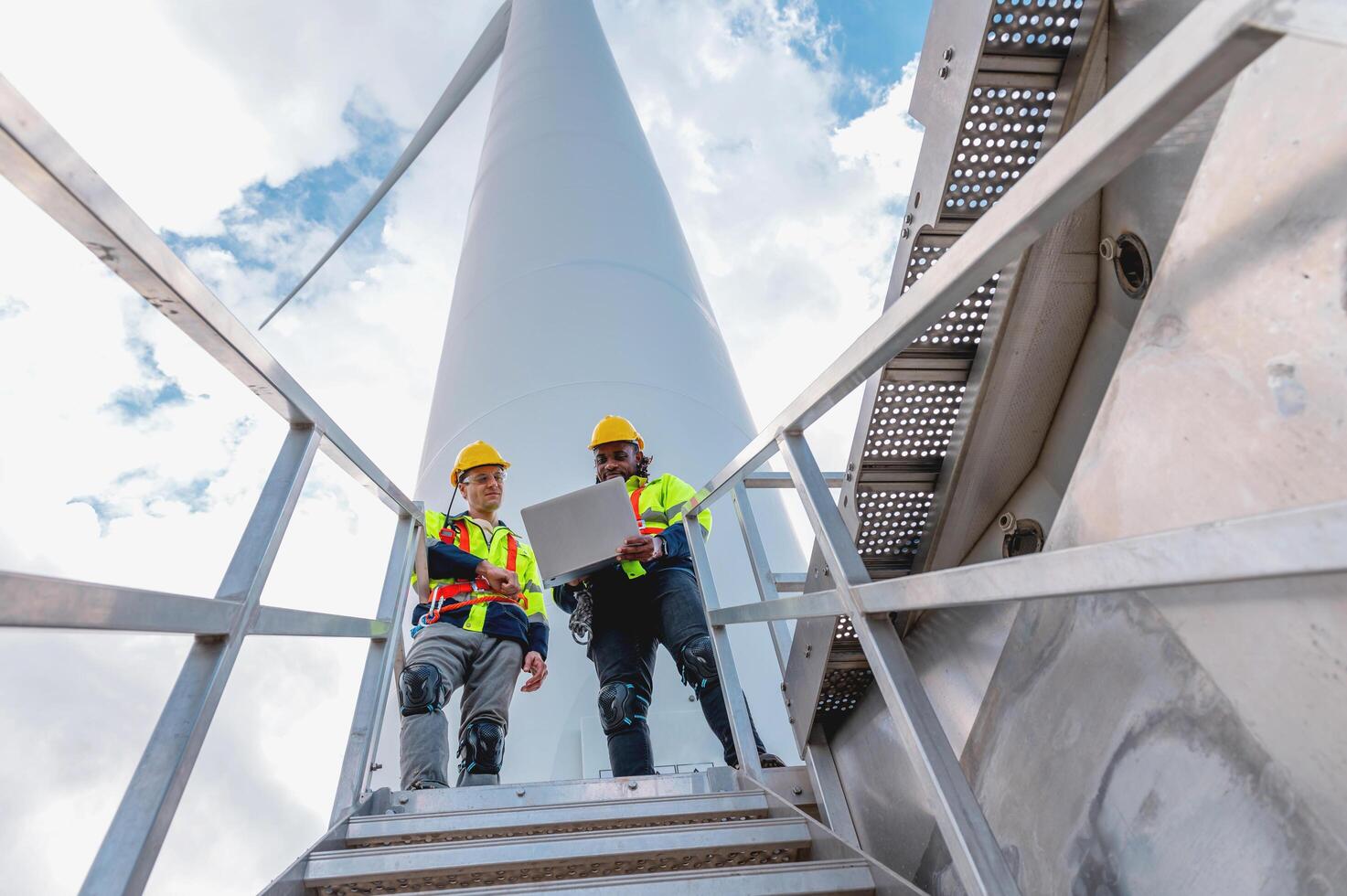 two men in safety gear are standing on a ladder next to a large wind turbine photo