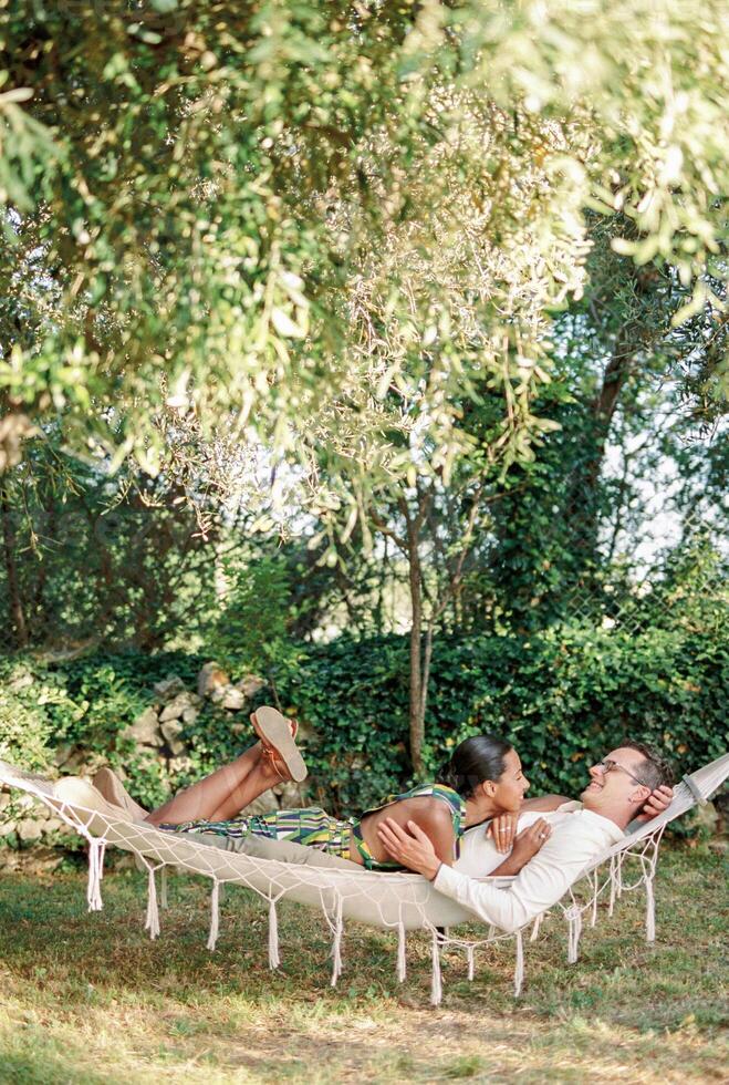 Woman resting her head on her arm on the chest of man hugging her waist while lying in a hammock in the garden photo