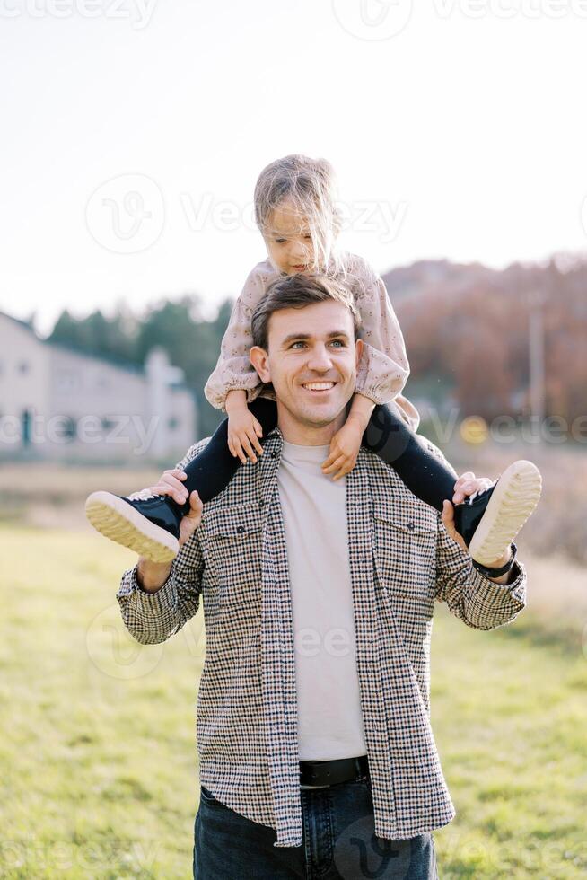 Smiling dad carries little girl on his shoulders across the meadow, looking to the side photo