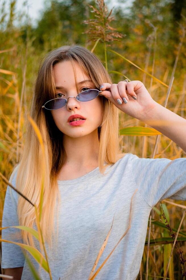 hermosa joven niña vistiendo blanco gris camiseta y negro pantalones en azul lentes posando en contra alto verde y amarillo césped en temprano calentar otoño. al aire libre retrato de hermosa hembra modelo. foto