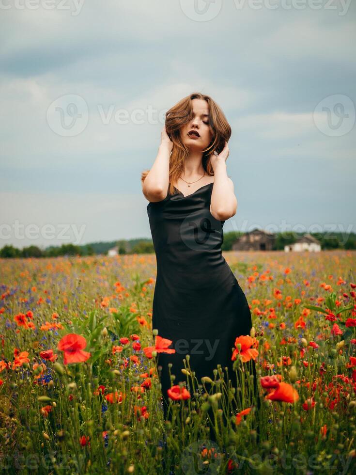 Beautiful young girl in a black evening dress posing against a poppy field on a cloudy summer day. Portrait of a female model outdoors. Rainy weather. Gray clouds. photo