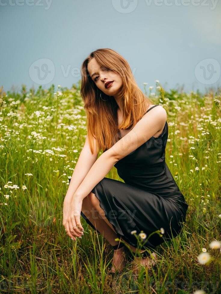 Beautiful young girl in a black evening dress poses crouching in front of a daisy field on a cloudy summer day. Portrait of a female model outdoors. Rainy weather. Gray clouds. photo