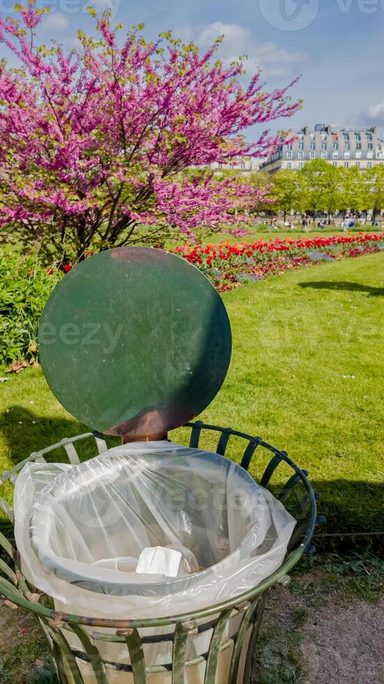 Public trash can with an open lid in a colorful spring park with blooming pink trees and flowers, conceptually related to Earth Day and environmental awareness photo