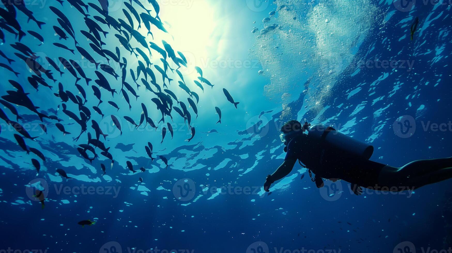 Underwater silhouette of a diver exploring a vibrant school of fish against the sunlit blue ocean, perfect for World Oceans Day and marine life conservation photo