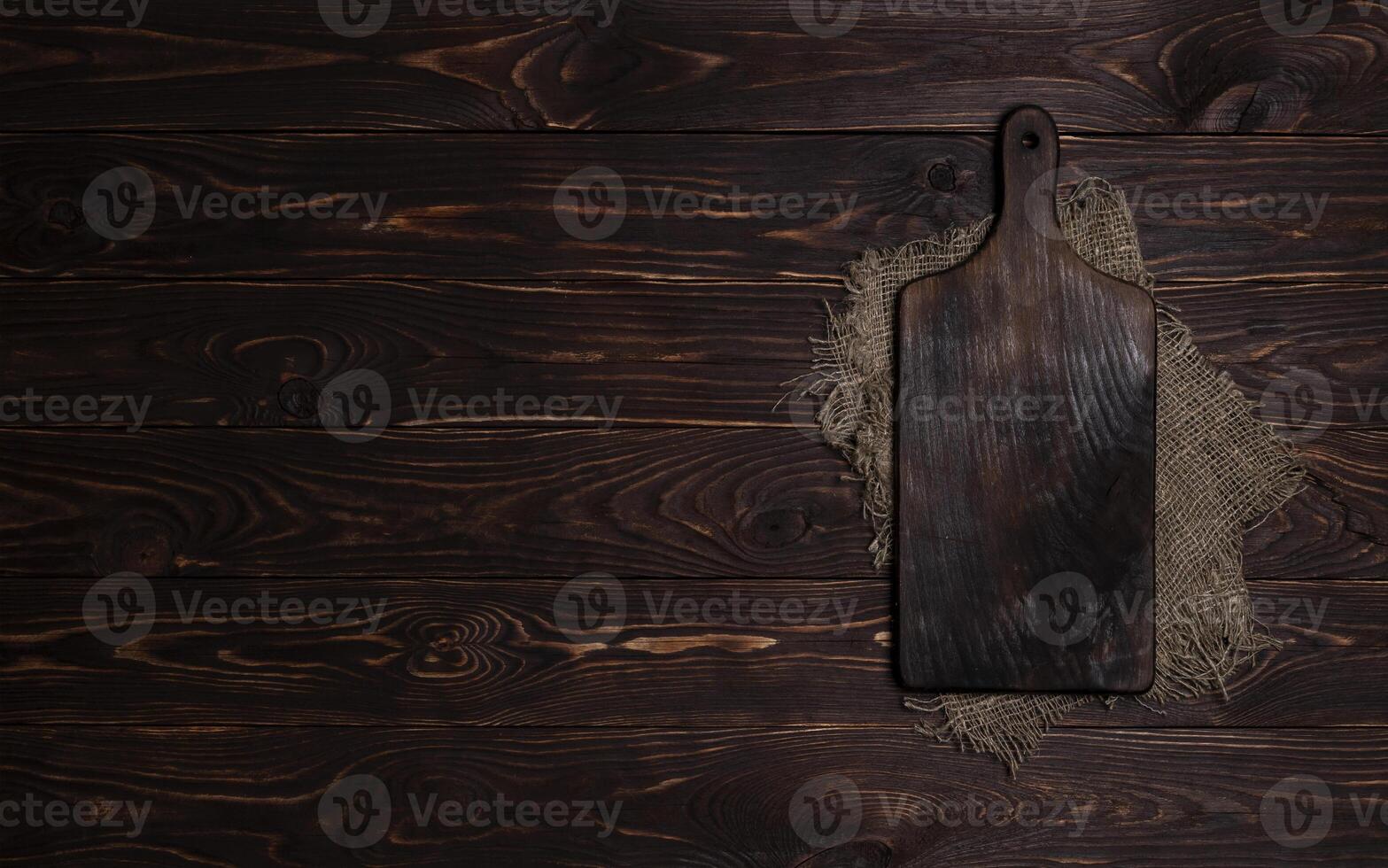 Cutting board with burlap napkin on brown wooden background, top view photo