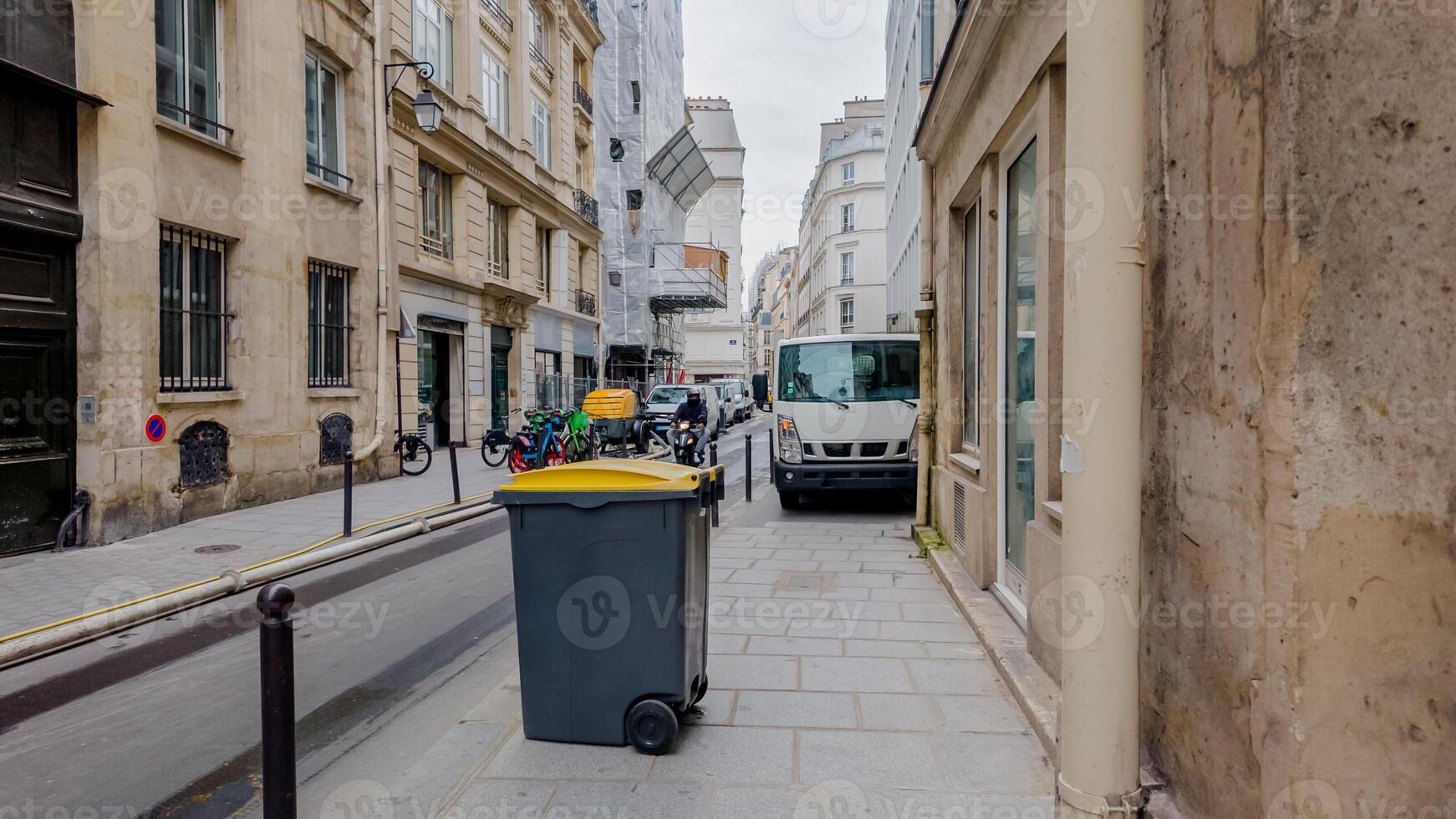 Quaint European city street with waste bins, bicycles, and parked vehicles, exemplifying urban life and municipal services on a cloudy day photo