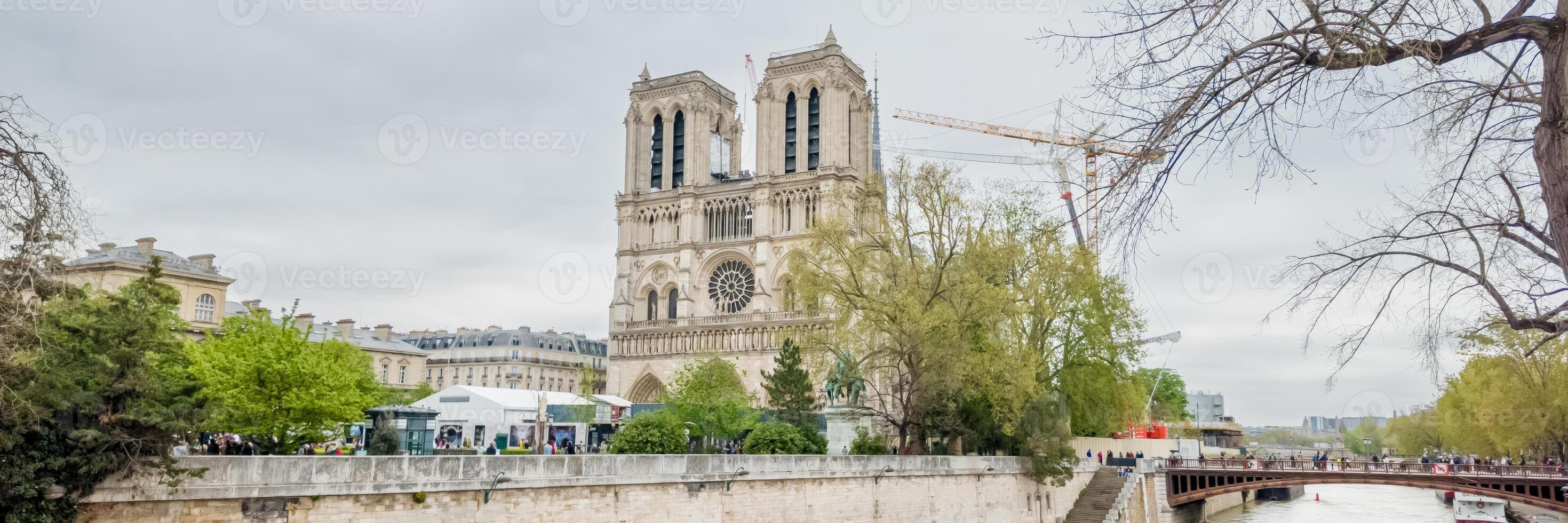 Panoramic view of the iconic Notre Dame Cathedral in Paris on a cloudy day, with surrounding spring foliage and a bustling riverwalk Related to, architecture, travel, heritage photo