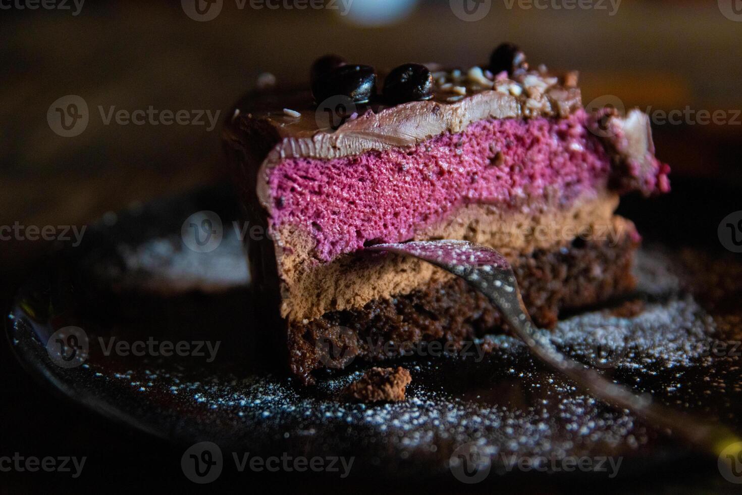 Piece of delicious homemade chocolate cake with blackberries and raspberries dusted with sugar powder served on the dark plate with fork on the wooden chess board table. Tasty Pastry ready for eating. photo
