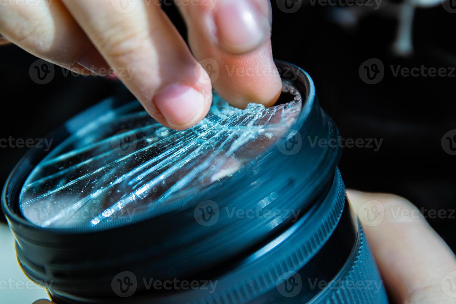 Photographer removing broken pieces of glass out of DSLR photo camera lens filter after if fall down onto the floor. Close up. Destroyed cracked glass of photo-filter. Macro. Top view. Selective focus