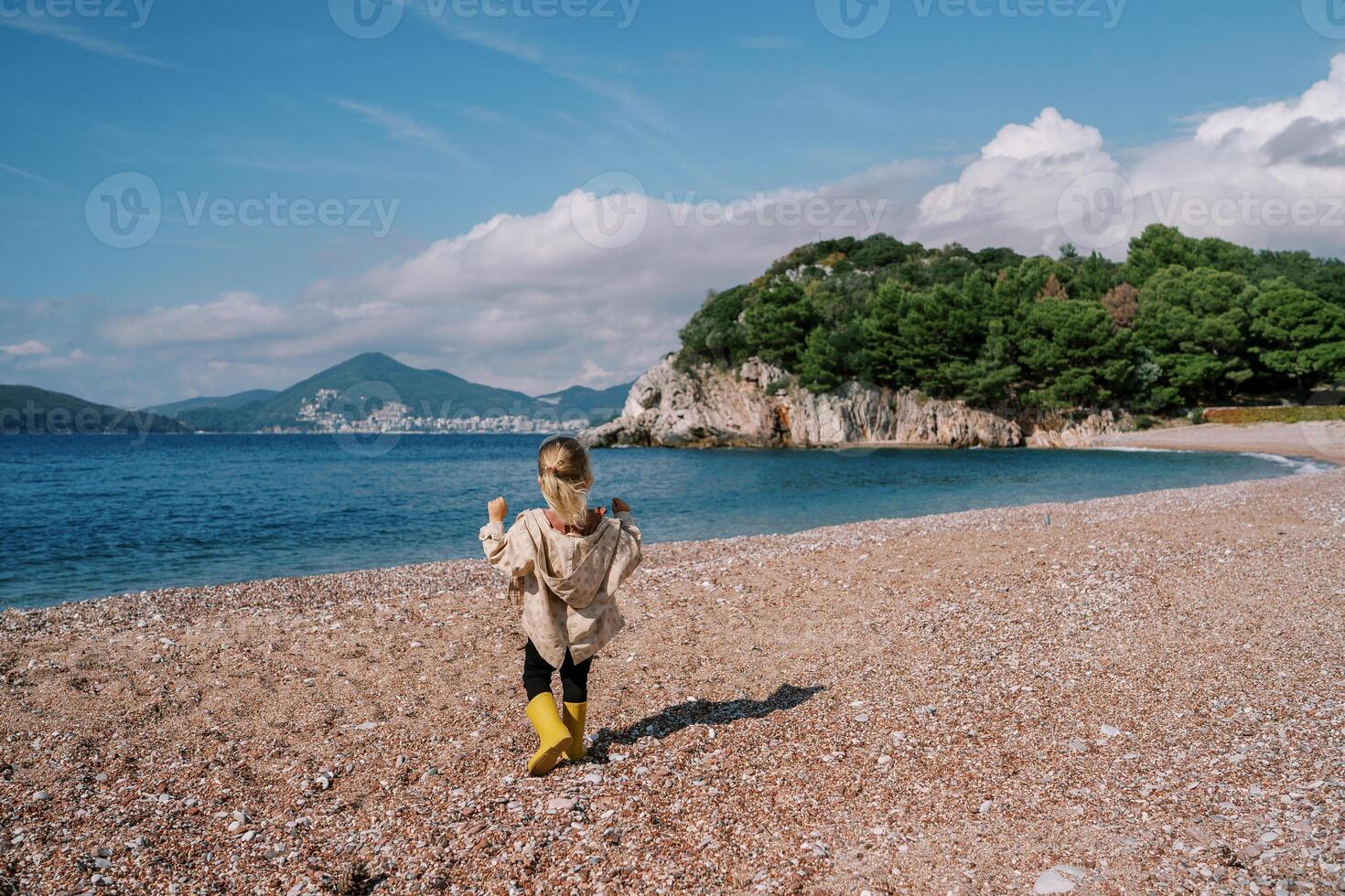 Little girl walks along the beach to the sea looking at the mountains. Back view photo