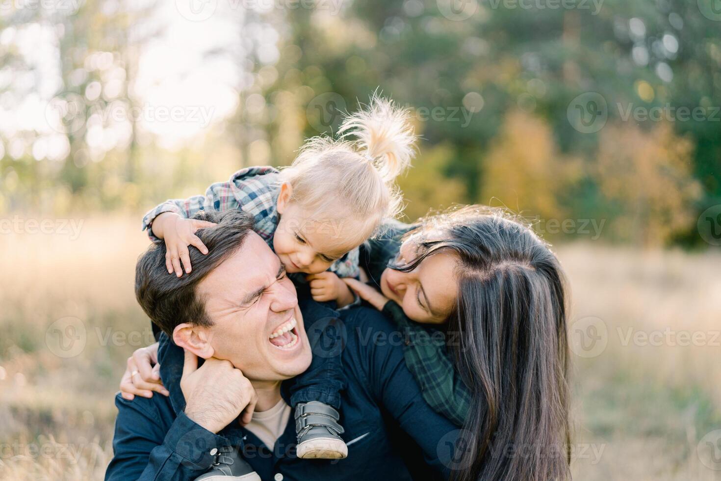 Laughing dad is sitting on the lawn with a little girl on his shoulders and mom hugging them from behind photo