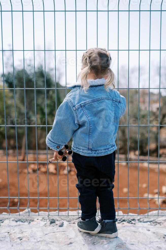 Little girl stands near a wire fence and looks through it at the park. Back view photo