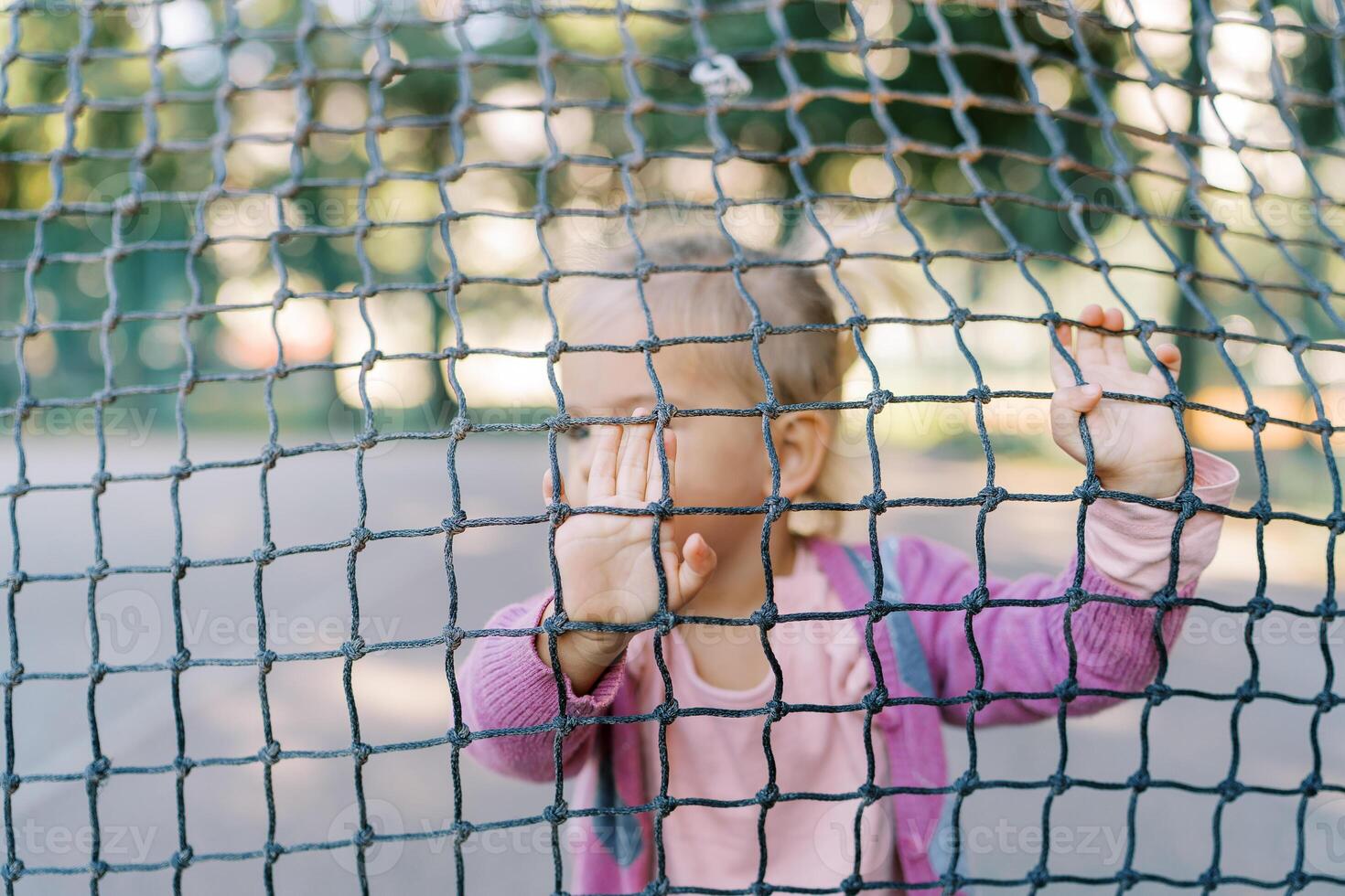 Little girl stands behind a tennis net and rests her hands on it photo