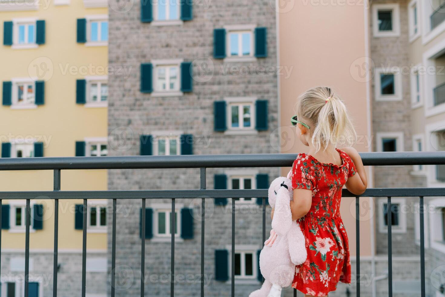 Little girl with a pink toy hare stands by the fence and looks at a colorful residential building. Back view photo