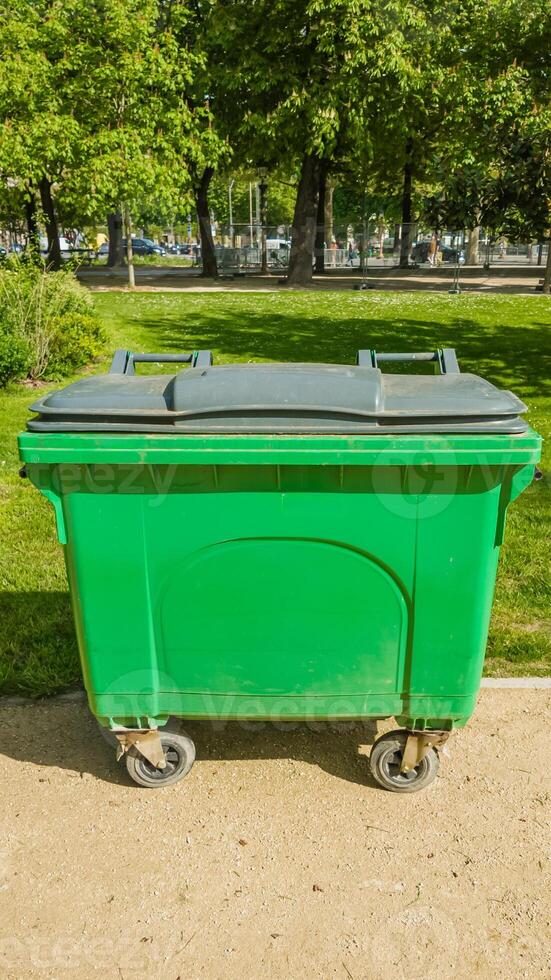 Large green commercial waste bin on wheels in a sunlit urban park setting, highlighting themes of environmental conservation and city sanitation photo