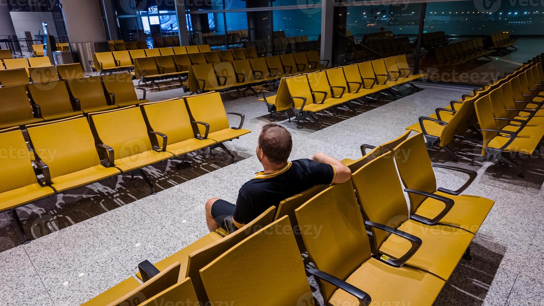 Adult man sitting alone amidst rows of empty chairs in a well lit airport waiting area, depicting travel, solitude, or potential layover scenarios photo