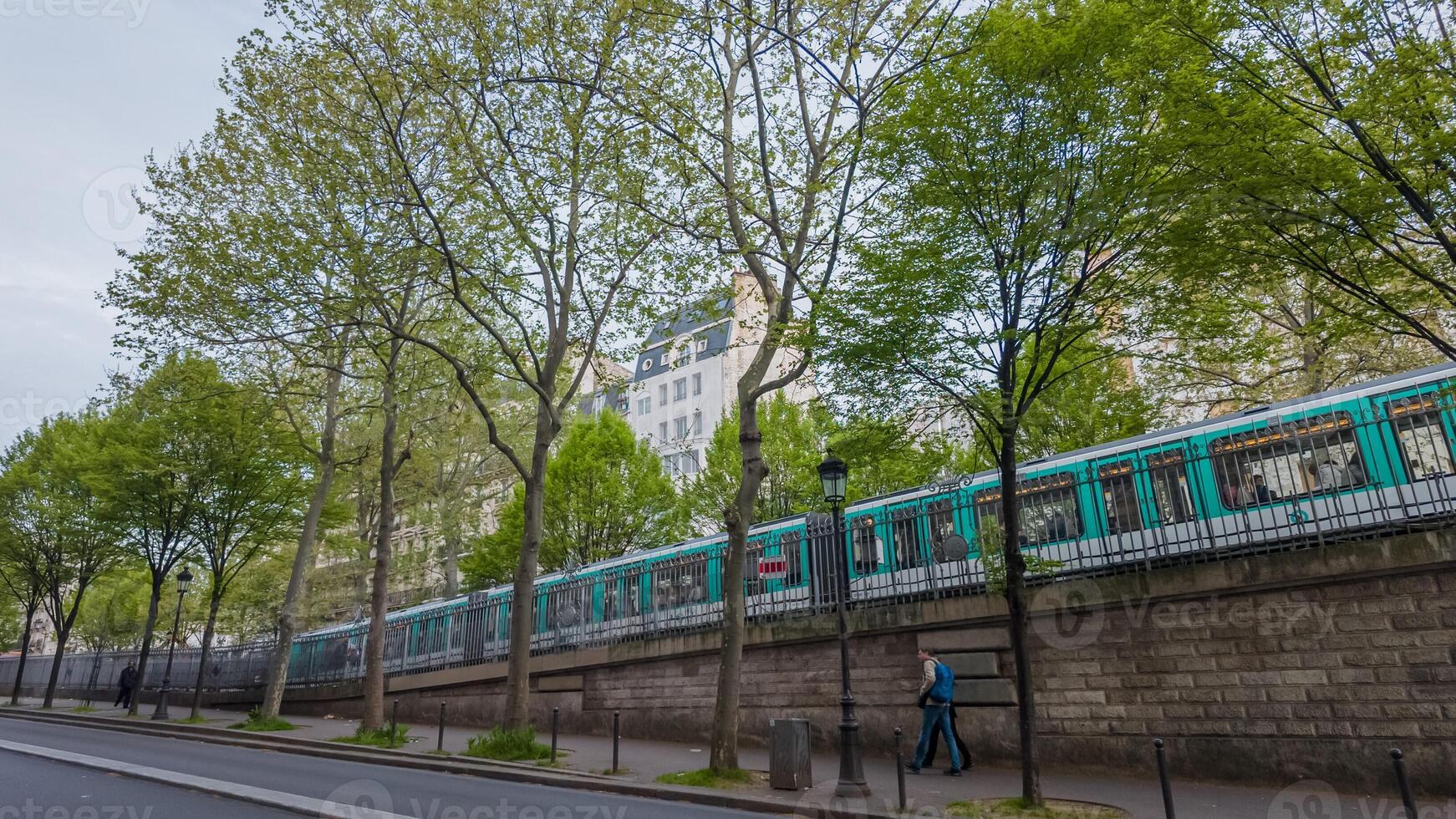 Pedestrian walking by a tree lined street with a passing metro train on an elevated track in a bustling city during springtime Related concepts, urban life, public transportation photo