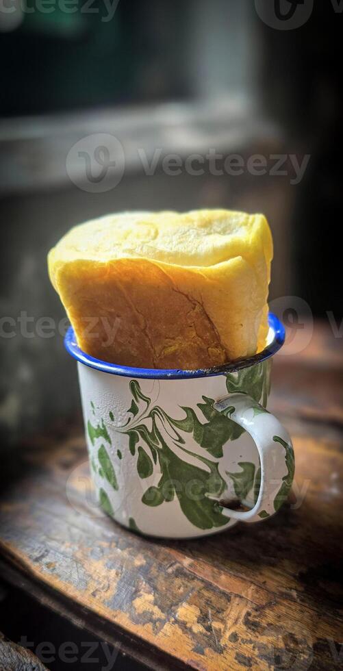 Ice chocolate drink on a vintage Indonesian iron mug with random green pattern called Blirik cup or cangkir Blirik on bokeh background along with chocolate filling bread photo