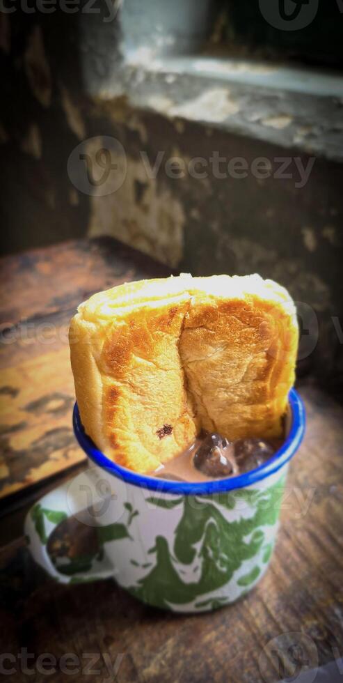 Ice chocolate drink on a vintage Indonesian iron mug with random green pattern called Blirik cup or cangkir Blirik on bokeh background along with chocolate filling bread photo