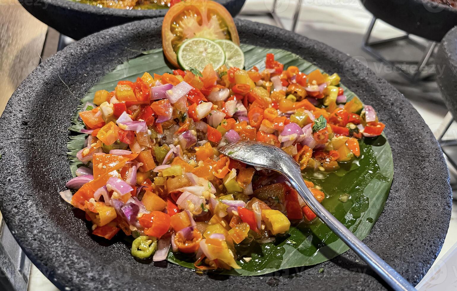 picante condimento de indonesio sambal Dabú Dabú hecho de chile, tomate y chalote con limón albahaca en un cerámico bol, de madera cuchara en gris antecedentes foto