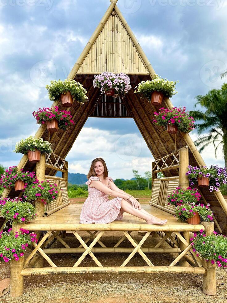 A woman in a pink floral dress stands beneath a striking bamboo archway adorned with vibrant flowers, set against a rural landscape, encapsulating the essence of rustic charm and natural beauty photo