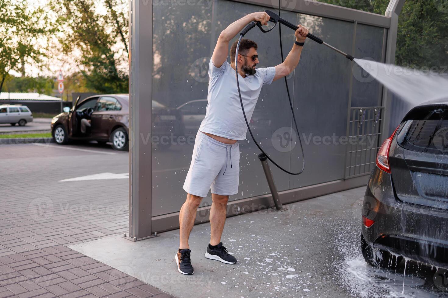 At a car wash, a man washes off the foam by lifting it over his head with a water cannon. A car at a self service car wash. photo