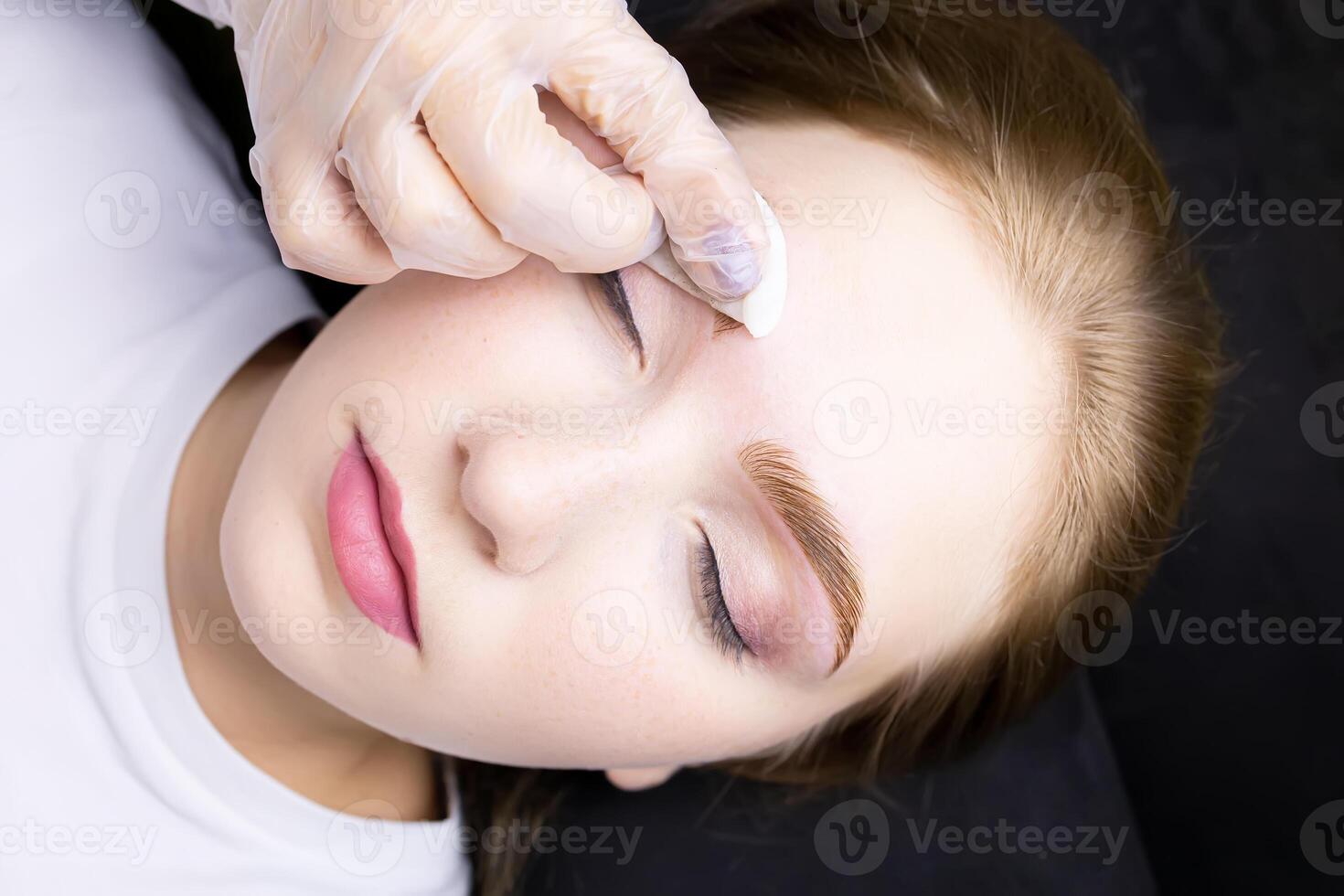 a close-up of the face of a blonde model who is lying on a black couch, the master wipes excess paint from her eyebrows with a cotton sponge photo