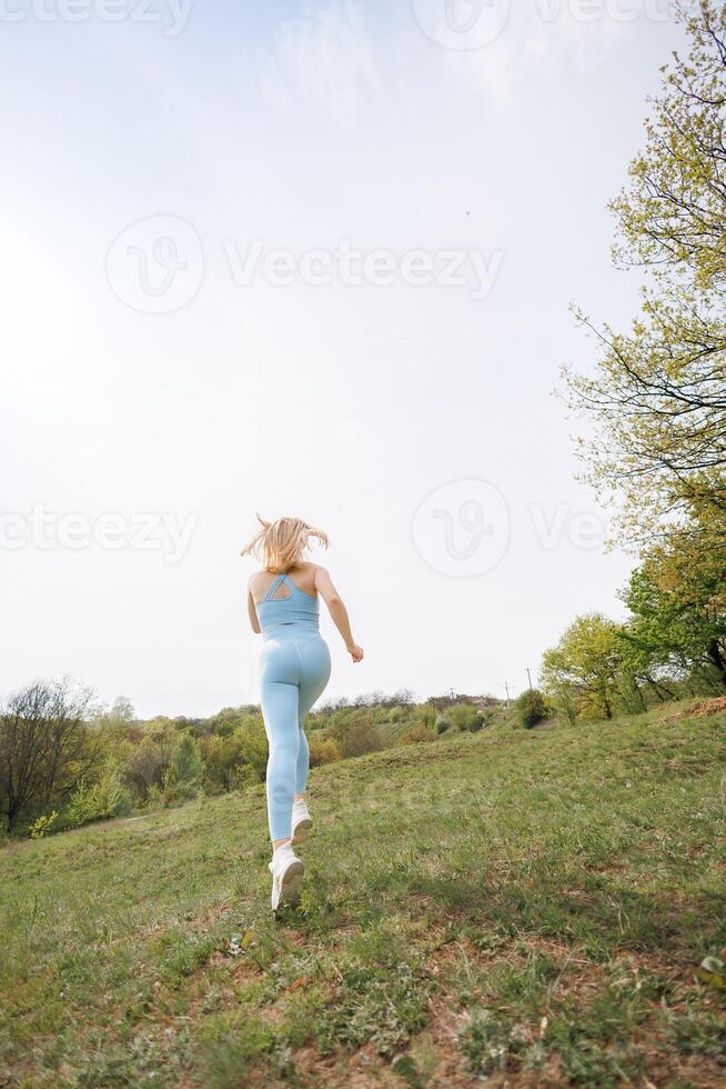 An athletic girl performs a jog in the park. Beautiful blonde Caucasian woman in blue tight tracksuit. Blonde girl at an outdoor training session photo