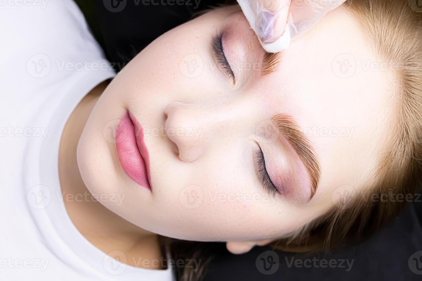 a close-up of the face of a blonde model who is lying on a black couch, the master wipes excess paint from her eyebrows with a cotton sponge photo