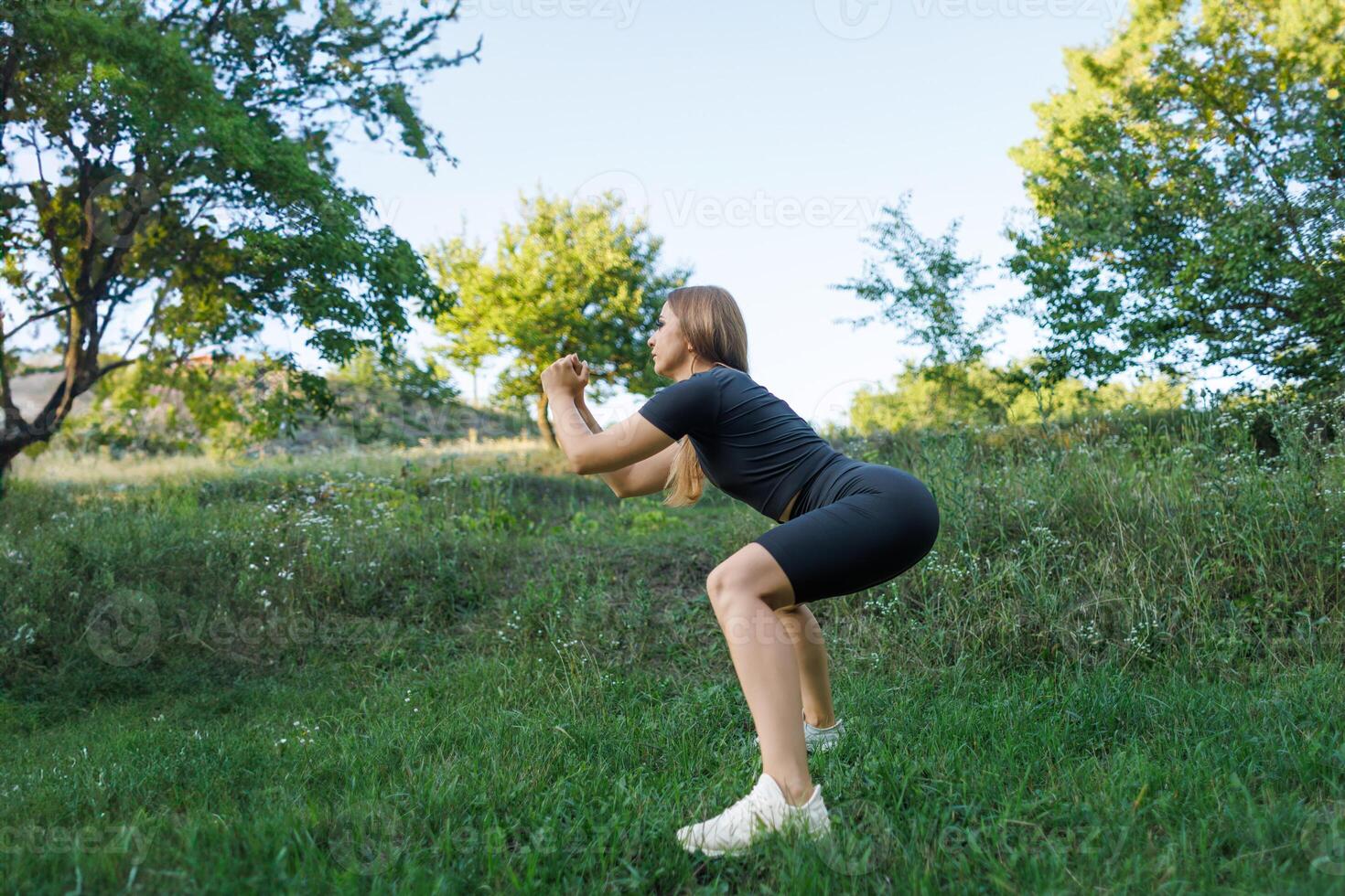 The girl performs athletic squats with her arms folded in front of her. Athletic girl in a tight uniform working outdoors in the park. photo