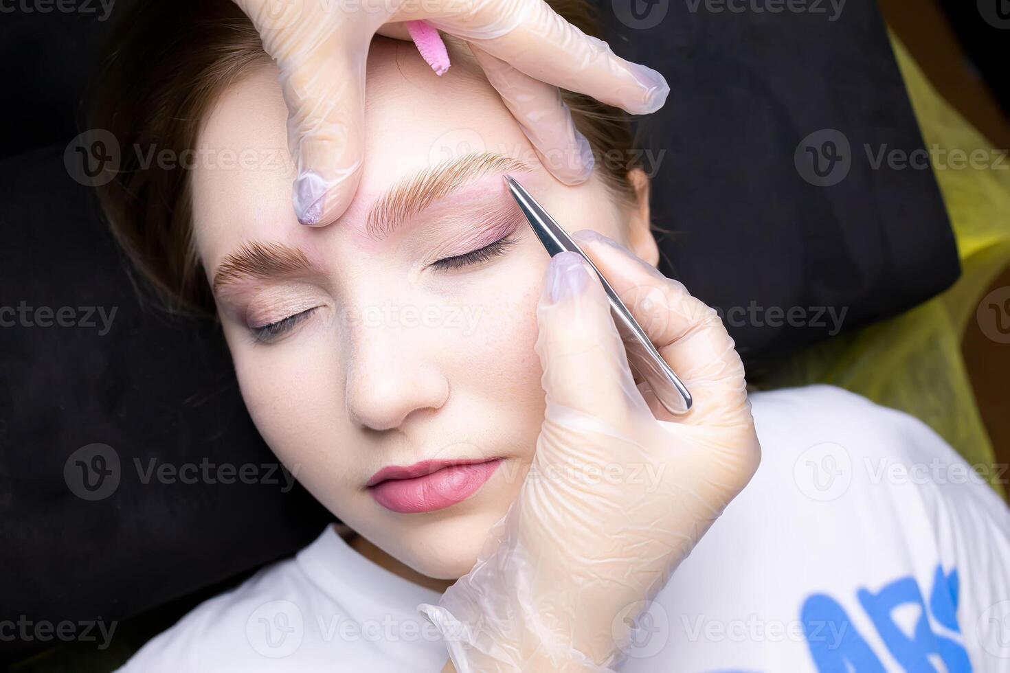 the master plucks out excess hairs with tweezers near the eyebrows after the procedure of long-term styling compositions photo