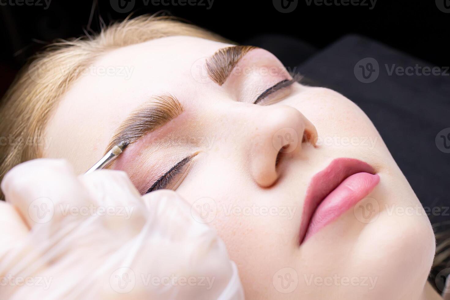 close-up of the eyebrows of the model on which the master applies a thick layer of paint for coloring the hairs of the eyebrows after lamination with compounds photo