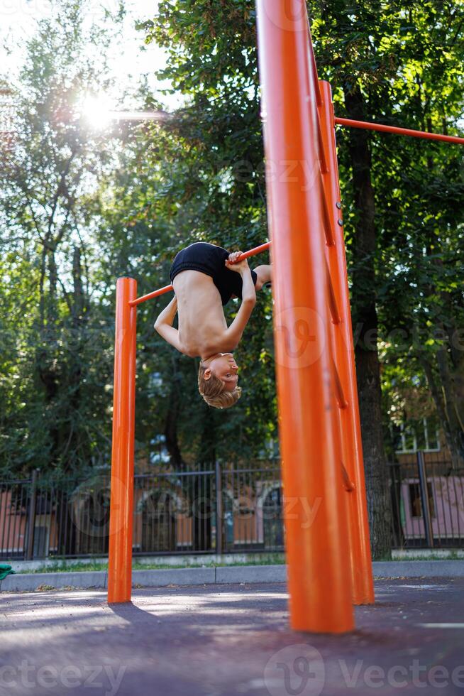 A teenager in the park goes in for sports hanging on the bar at the bottom with his head. Street workout on a horizontal bar in the school park. photo