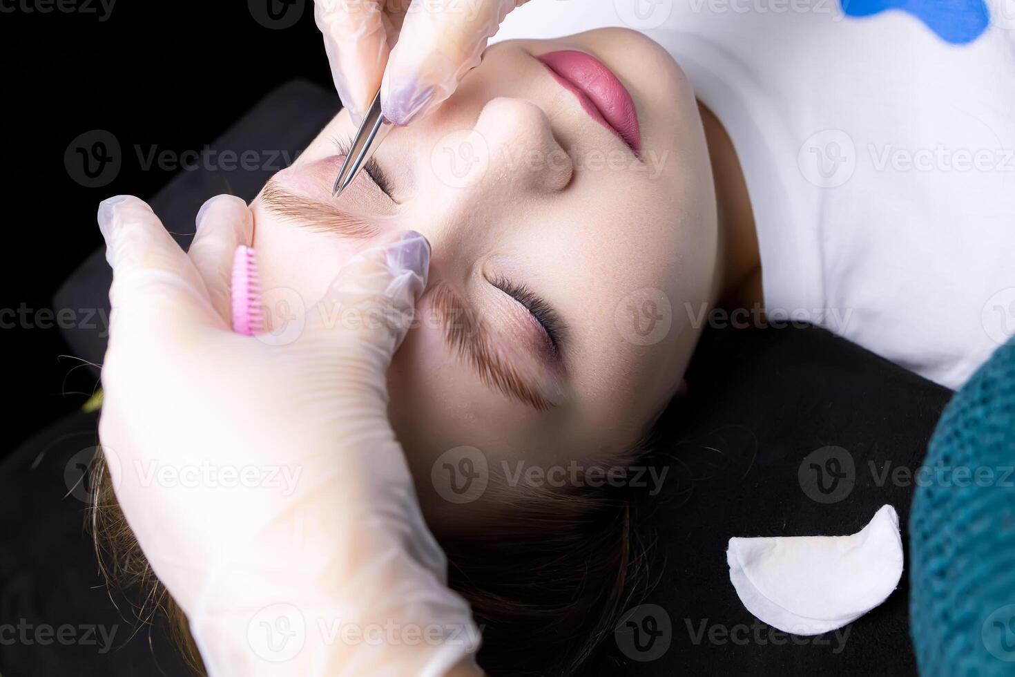 the hands of the master in white gloves perform plucking of excess hairs with tweezers after laminating the eyebrows photo