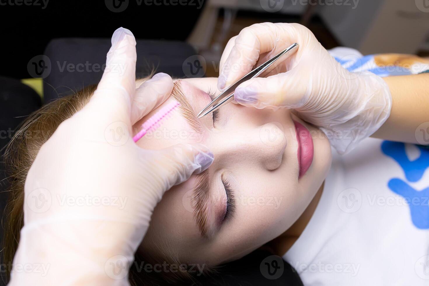 the master's hands in white gloves stretch the eyebrow and pluck out excess eyebrow hairs with tweezers photo