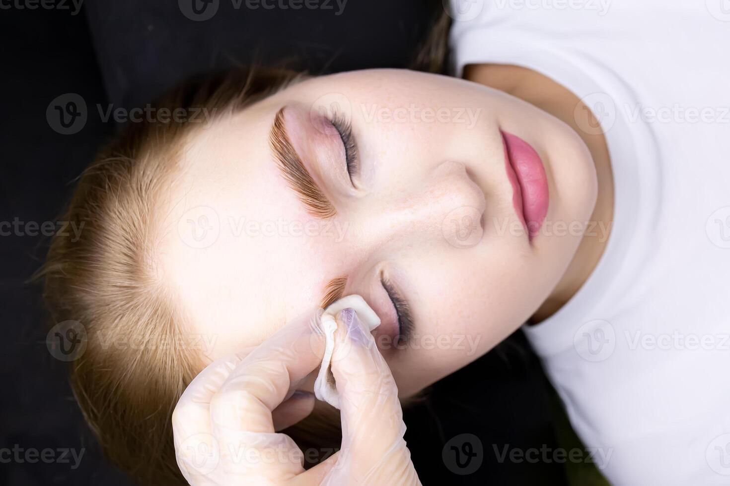 a close-up of the face of a blonde model who is lying on a black couch, the master wipes excess paint from her eyebrows with a cotton sponge photo