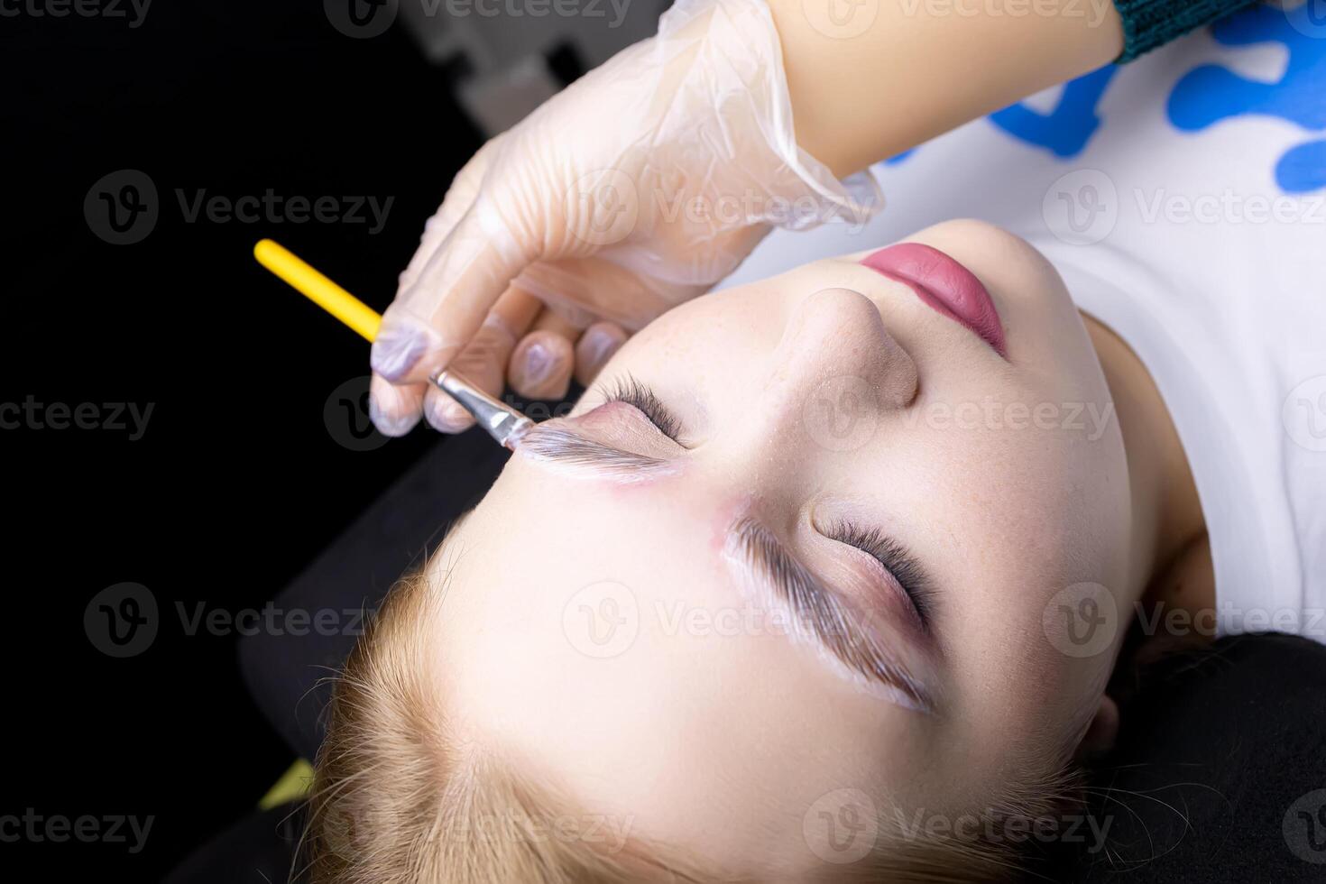 the blonde model is lying on a black couch with a white T-shirt, she is undergoing the procedure of lamination of eyebrows and photo