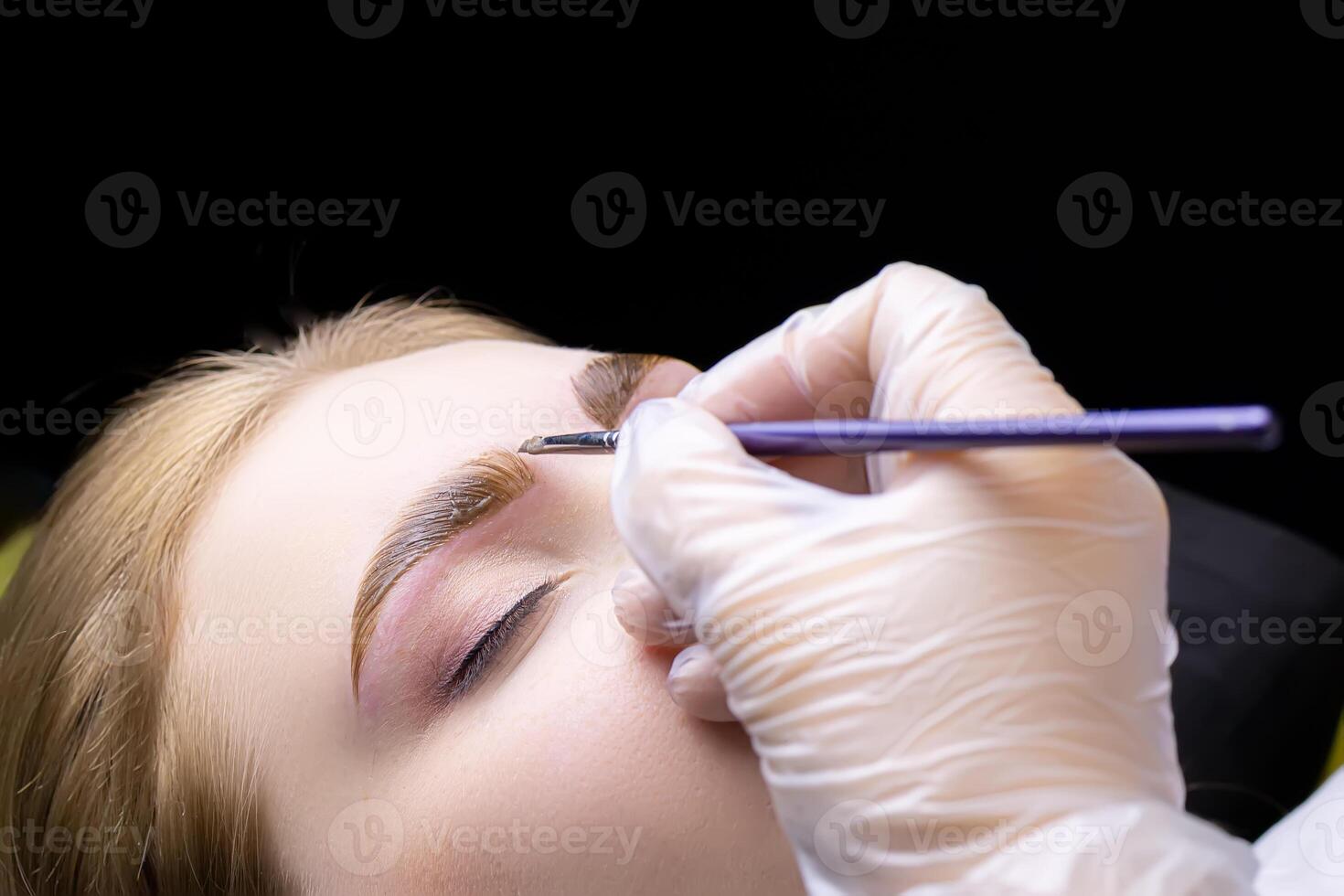 close-up of the eyebrows of the model on which the master applies a thick layer of paint for coloring the hairs of the eyebrows after lamination with compounds photo