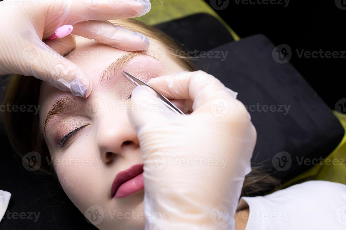 the master plucks out excess hairs with tweezers near the eyebrows after the procedure of long-term styling compositions photo