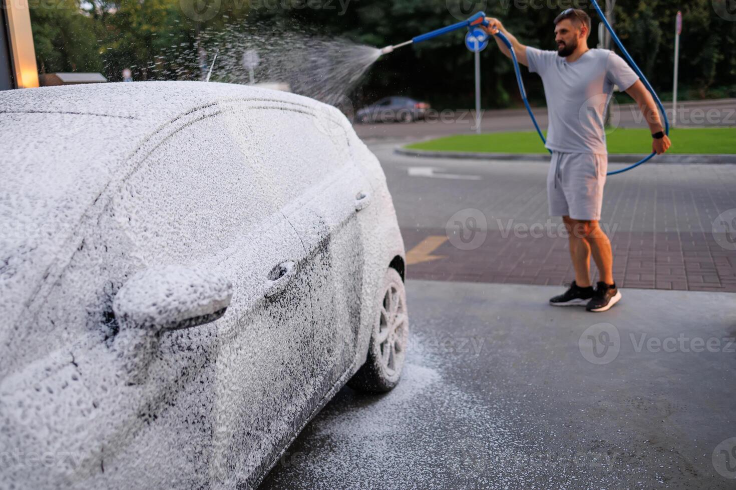 Side view of a car covered in foam, man in the background applying foam to the car. A car at a self service car wash. photo