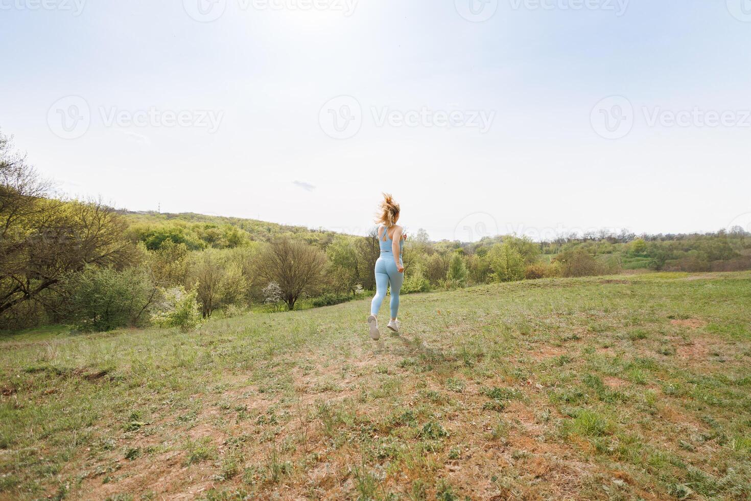 An athlete performs running drills in the park. Beautiful blonde Caucasian woman in blue tight tracksuit. Blonde girl at an outdoor training session photo