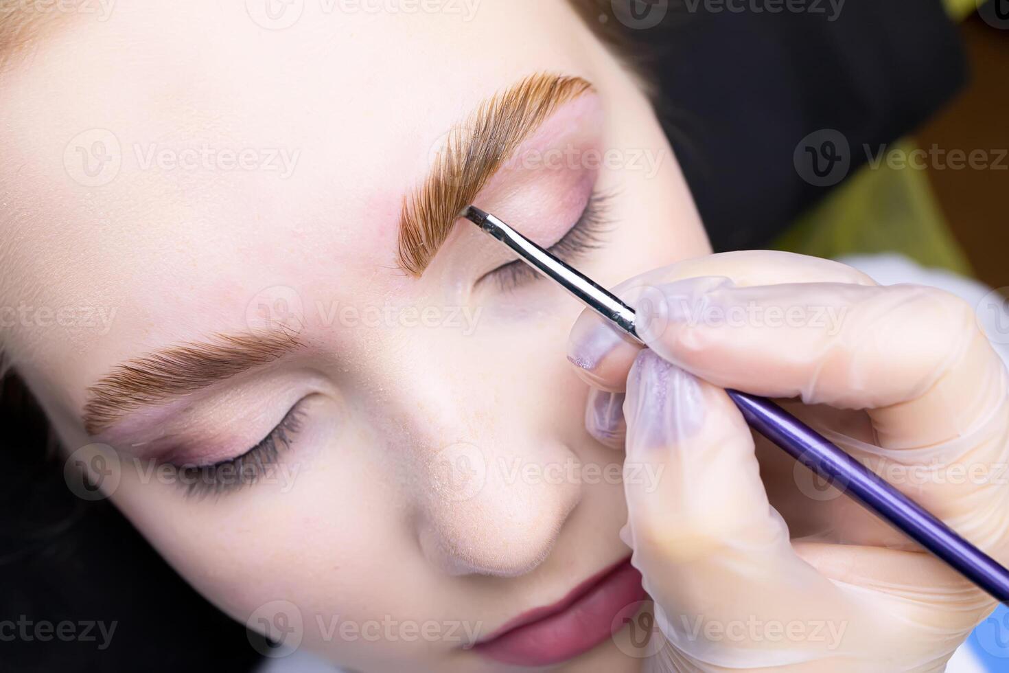 the hands of the master hold a brush and apply paint to the eyebrows of the model coloring the hairs after the lamination procedure photo